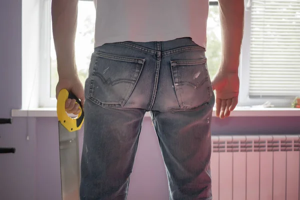 Человек с ручной пилой в старых грязных джинсах стоит у окна Стоковая Картинка