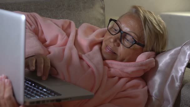 Kadın kıdemli bir ev iç kanepede yatan bir dizüstü bilgisayar kullanarak. Yakın çekim — Stok video