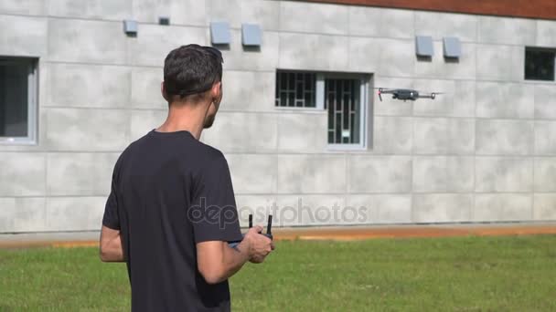Чоловік контролює безпілотник біля сірої будівлі на зеленому газоні. Літо. Крупним планом — стокове відео