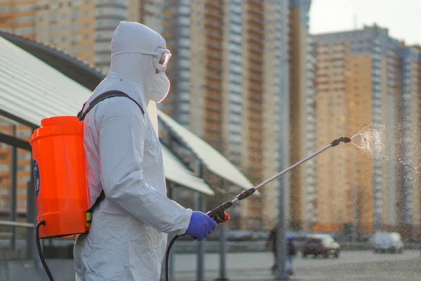 Koruyucu ekipman kullanan bir adam şehirde dezenfektanla dezenfekte ediyor. Coronavirus covid-19 hastalığından dolayı yüzey tedavisi. Beyaz takım elbiseli bir adam elinde sprey silahla caddeyi dezenfekte ediyor. Virüs salgını