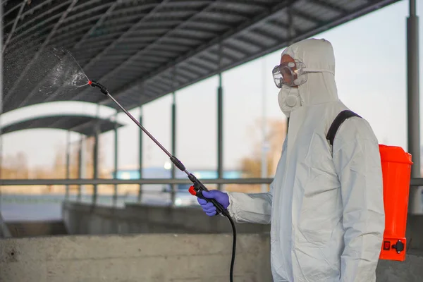 Koruyucu ekipman kullanan bir adam şehirde dezenfektanla dezenfekte ediyor. Coronavirus covid-19 hastalığından dolayı yüzey tedavisi. Beyaz takım elbiseli bir adam elinde sprey silahla caddeyi dezenfekte ediyor. Virüs salgını