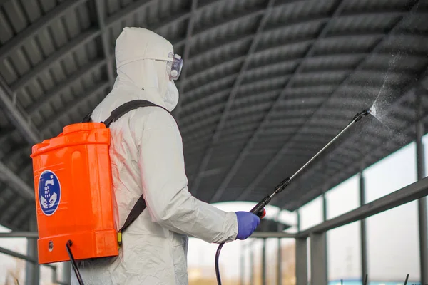 Koruyucu ekipman kullanan bir adam şehirde dezenfektanla dezenfekte ediyor. Coronavirus covid-19 hastalığından dolayı yüzey tedavisi. Beyaz takım elbiseli bir adam elinde sprey silahla caddeyi dezenfekte ediyor. Virüs salgını