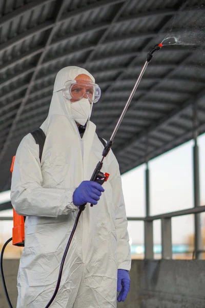 Koruyucu ekipman kullanan bir adam şehirde dezenfektanla dezenfekte ediyor. Coronavirus covid-19 hastalığından dolayı yüzey tedavisi. Beyaz takım elbiseli bir adam elinde sprey silahla caddeyi dezenfekte ediyor. Virüs salgını