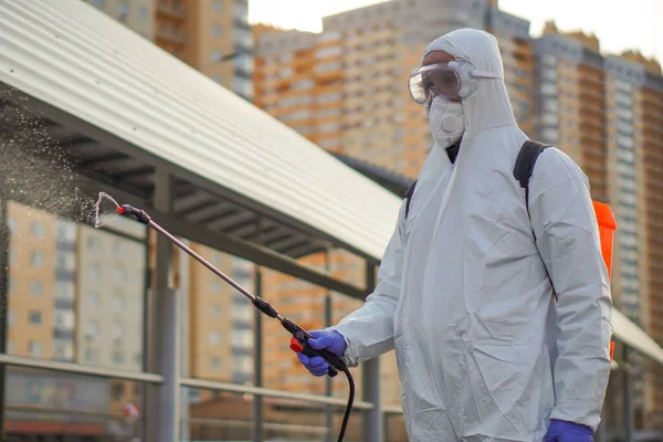 Koruyucu ekipman kullanan bir adam şehirde dezenfektanla dezenfekte ediyor. Coronavirus covid-19 hastalığından dolayı yüzey tedavisi. Beyaz takım elbiseli bir adam elinde sprey silahla caddeyi dezenfekte ediyor. Virüs salgını