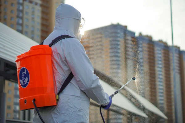 Koruyucu ekipman kullanan bir adam şehirde dezenfektanla dezenfekte ediyor. Coronavirus covid-19 hastalığından dolayı yüzey tedavisi. Beyaz takım elbiseli bir adam elinde sprey silahla caddeyi dezenfekte ediyor. Virüs salgını