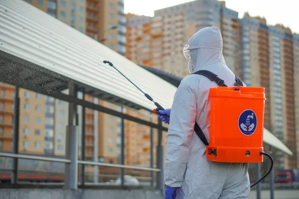 Koruyucu ekipman kullanan bir adam şehirde dezenfektanla dezenfekte ediyor. Coronavirus covid-19 hastalığından dolayı yüzey tedavisi. Beyaz takım elbiseli bir adam elinde sprey silahla caddeyi dezenfekte ediyor. Virüs salgını