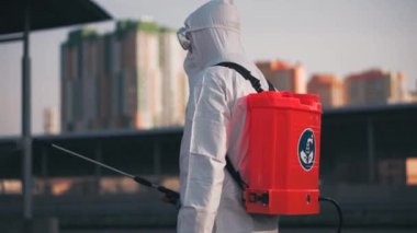 Koruyucu ekipman kullanan bir adam şehirde dezenfektanla dezenfekte ediyor. Coronavirus covid-19 hastalığından dolayı yüzey tedavisi. Beyaz takım elbiseli bir adam elinde sprey silahla caddeyi dezenfekte ediyor. Virüs salgını