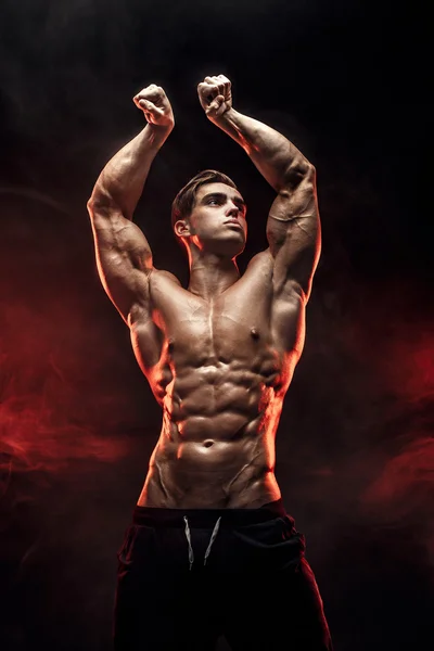 Hombre fuerte culturista con abdominales perfectos, hombros, bíceps, tríceps, pecho posando en humo manos arriba . — Foto de Stock