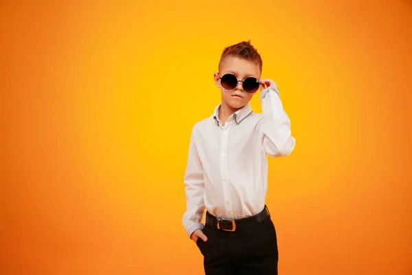 Elegante niño con camisa blanca y pantalones negros posando en gafas de sol sobre fondo naranja . — Foto de Stock