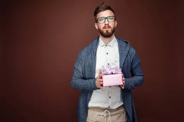 Junger Mann mit Brille und lässigem Outfit hält kleine rosa Geschenkschachtel auf braunem Studiohintergrund. — Stockfoto