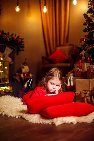 Petite fille couchée sur le tapis avec des cadeaux autour en utilisant une tablette sur un oreiller rouge . — Photo