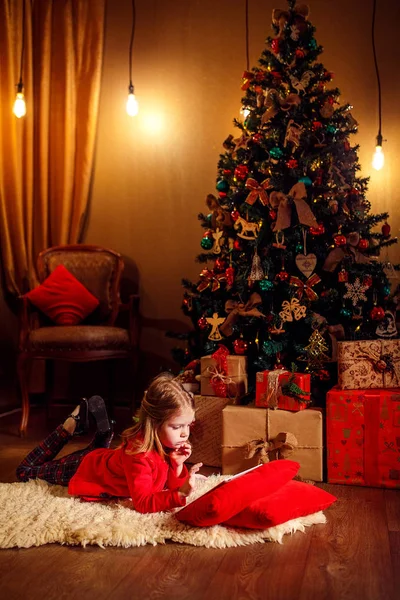 Petite fille couchée sur le tapis avec des cadeaux autour en utilisant une tablette sur un oreiller rouge . — Photo