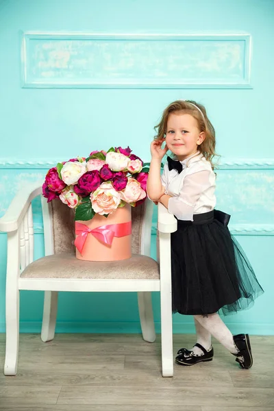 Kleines Mädchen in weißem Hemd, schwarzem Rock und Fliege posiert lächelnd in die Kamera neben Stuhl, blaue Stuckwand. — Stockfoto