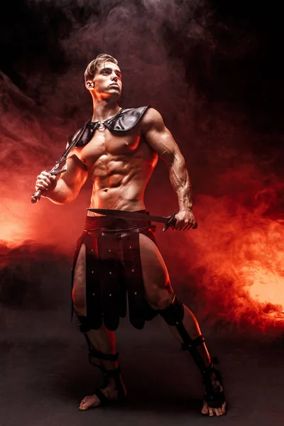 Retrato de larga duración de un joven guerrero sexy sosteniendo la espada y mirando hacia otro lado mientras posa contra el fuego. Espacio de copia — Foto de Stock