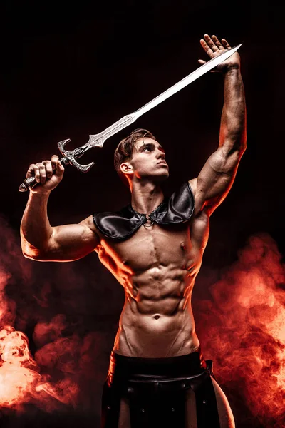 Young muscular model posing with sword in hands and looking away in fire background. — Stock Photo, Image