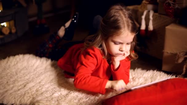 Petite fille couchée sur le tapis avec des cadeaux autour en utilisant la tablette sur — Video