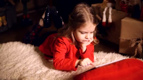 Niña acostada en la alfombra con regalos alrededor usando tableta en — Vídeo de stock