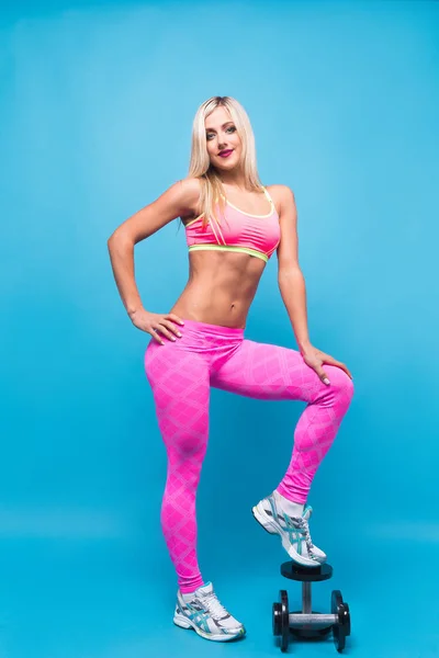 Retrato de mujer rubia atractiva en ropa deportiva rosa posando con mancuernas sobre fondo azul —  Fotos de Stock