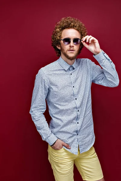 Stijlvolle curly-haired mens in zonnebril op rode achtergrond — Stockfoto