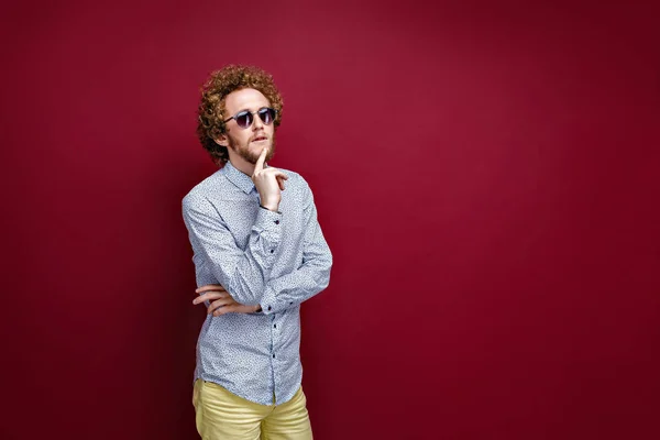 Stylish curly-haired man in sunglasses on red background. Thinki — Stock Photo, Image