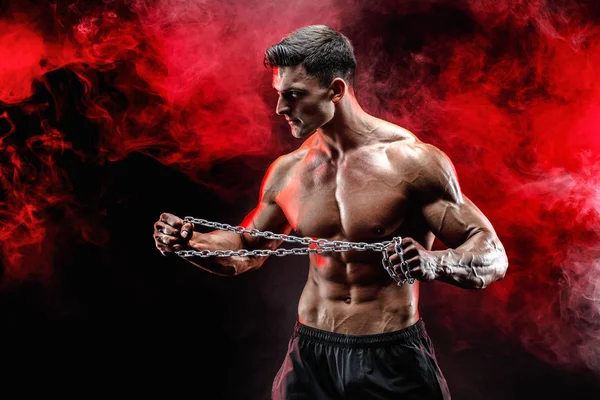 金属チェーンを引き裂く筋肉スポーツマンの肖像画。赤い煙が黒の背景 — ストック写真