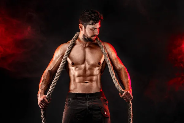 М'язистий чоловік тримає мотузку і позує на чорному тлі з червоним димом — стокове фото