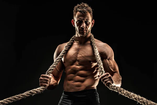 En stilig sexuella stark ung man med muskulös kropp håller repet med händer hänger på nacke och axlar stående poserar i studio på svart bakgrund — Stockfoto