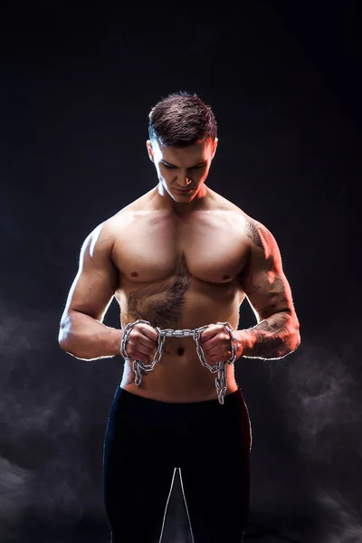 Knappe macht atletische man bodybuilder doen oefeningen met ketting, scheuren. Fitness gespierd lichaam op donkere achtergrond. Perfecte man. Awesome bodybuilder, tatoeage, poseren. — Stockfoto