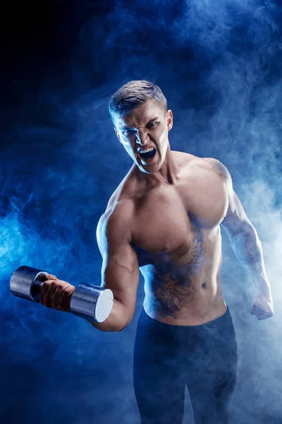 Close-up van een knappe macht atletische man bodybuilder doen oefeningen met dumbbell. Fitness gespierd lichaam op donkere rook achtergrond. Perfecte man. Awesome bodybuilder, tatoeage, poseren. — Stockfoto