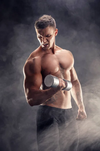 Close-up van een knappe macht atletische man bodybuilder doen oefeningen met dumbbell. Fitness gespierd lichaam op donkere rook achtergrond. Perfecte man. Awesome bodybuilder, tatoeage, poseren. — Stockfoto