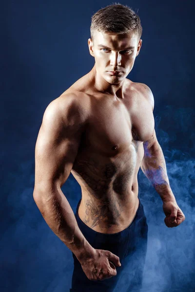 Hermoso poder atlético hombre culturista. Cuerpo muscular fitness sobre fondo de humo oscuro. Hombre perfecto. Culturista impresionante, tatuaje, posando . — Foto de Stock