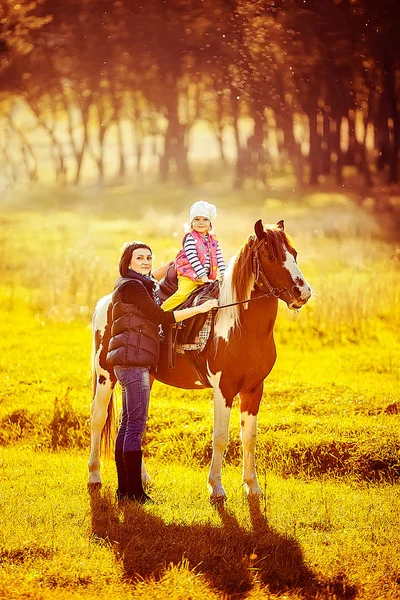 Liten flicka som rider på en häst med mamman promenader i närheten. — Stockfoto