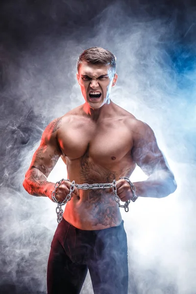 Portrait of muscular tattooed sportsman with metal chain. Background with smoke