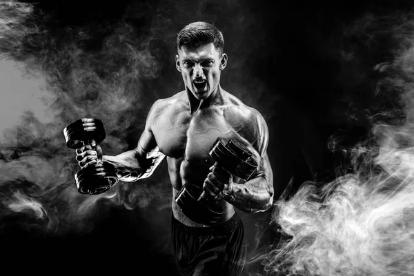 Handsome bodybuilder doing exercise with dumbbell. Studio shot. Black and white photo. Smoke — Stock Photo, Image