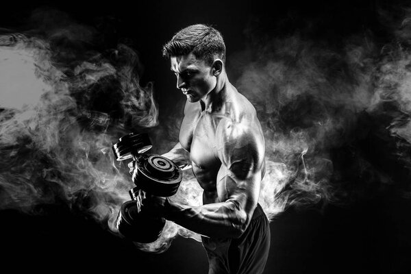 Handsome bodybuilder doing exercise with dumbbell. Studio shot. Black and white photo. Smoke