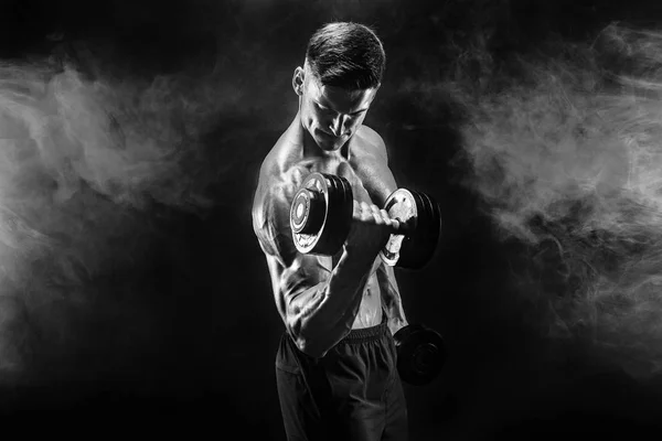 Guapo culturista haciendo ejercicio con mancuerna. Grabado en estudio. Foto en blanco y negro. Humo. — Foto de Stock