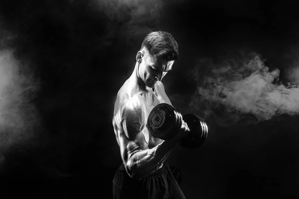 Handsome bodybuilder doing exercise with dumbbell. Studio shot. Black and white photo. Smoke — Stock Photo, Image