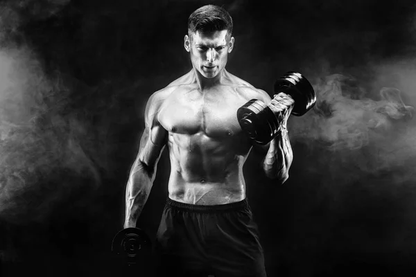 Handsome bodybuilder doing exercise with dumbbell. Studio shot. Black and white photo. Smoke — Stock Photo, Image