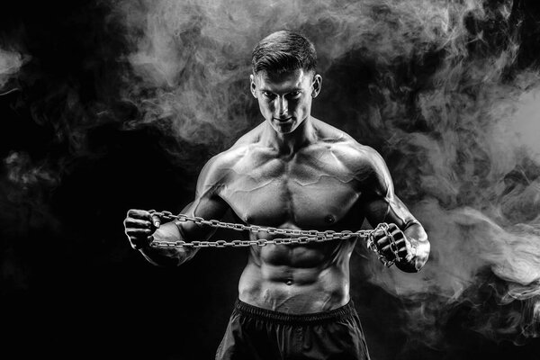 Portrait of muscular sportsman tearing metal chain.Black background with smoke