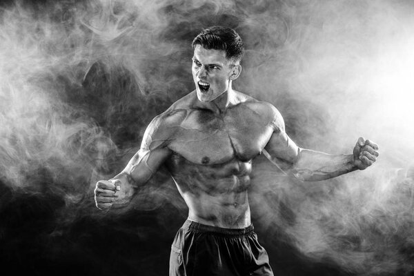Strong bodybuilder man with perfect body- abs, shoulders, biceps, triceps, chest posing in studio smoke background