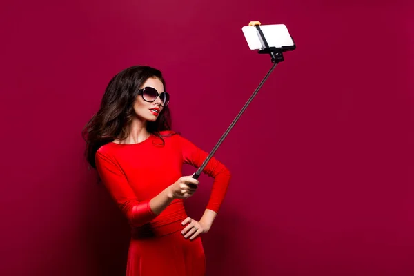 Portret van prachtige brunette in geweldige jurk en red lips dragen zonnebril terwijl het maken van de selfie met stok. Geïsoleerd — Stockfoto