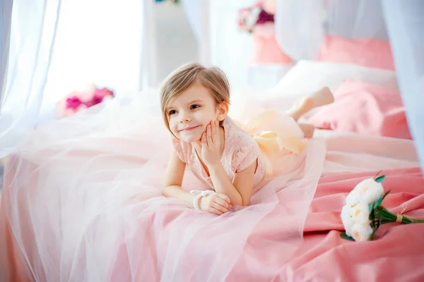 Kleines nettes Mädchen im Kleid, das auf dem Bett mit Blumen liegt — Stockfoto