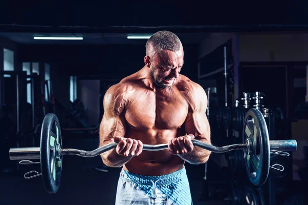 Muscular fisiculturista cara fazendo exercícios em bíceps com grande haltere no ginásio — Fotografia de Stock