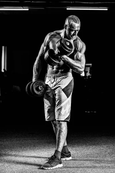 Retrato de um jovem no ginásio, exercício para o bíceps. Preto e branco. Inflação da lente. Baixa luz — Fotografia de Stock