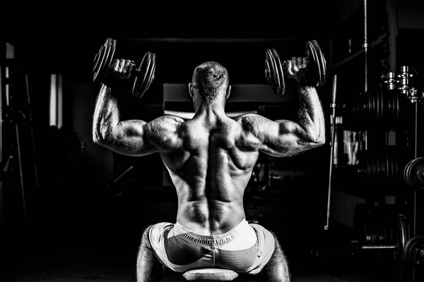 Homem musculoso treinando seus ombros com halteres no ginásio — Fotografia de Stock