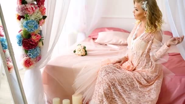 Junge blonde hübsche Frau in romantischem Negligee, die auf einem mit Blumen dekorierten Bett sitzt und sich selbst im Spiegel betrachtet. — Stockvideo