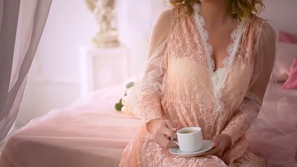 Giovane bionda bella donna in negligee romantico bere caffè mentre seduto su un letto decorato con fiori . — Video Stock