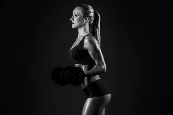 Side view of sexual muscular blonde with ponytail in bikini posing with dumbbells on black background. — Stock Photo, Image