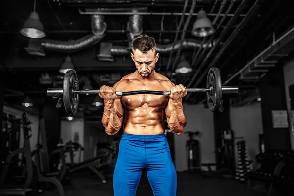 Joven apuesto culturista caucásico deportista haciendo ejercicio con una barra en el gimnasio, haciendo el entrenamiento de bíceps . — Foto de Stock