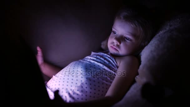 Маленька мила дівчинка сидить на дивані в повній темряві і грає з планшетом — стокове відео